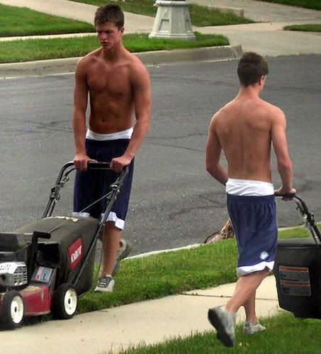 two shirtless young man wearing baggy shorts are mowing a lawn a suburban street